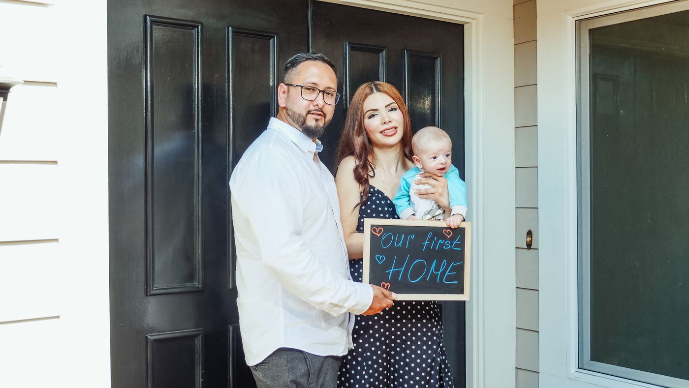 A young family holding an 'Our first home' sign at their front door